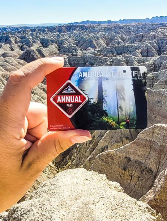 A hand holding the 2022 national park pass in front of Badlands National Park in South Dakota