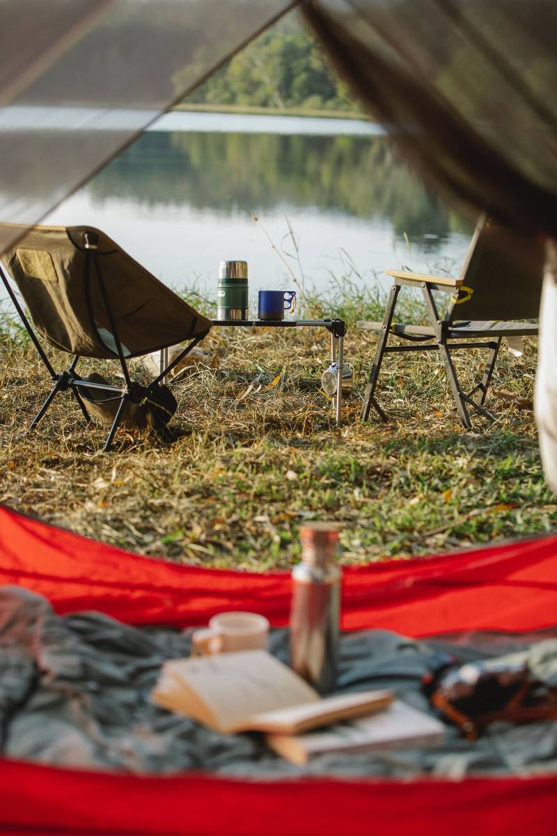 Two chairs near a folding table with a coffee cup and thermos sitting on top, all camping must haves, and a tent and book in the foreground.
