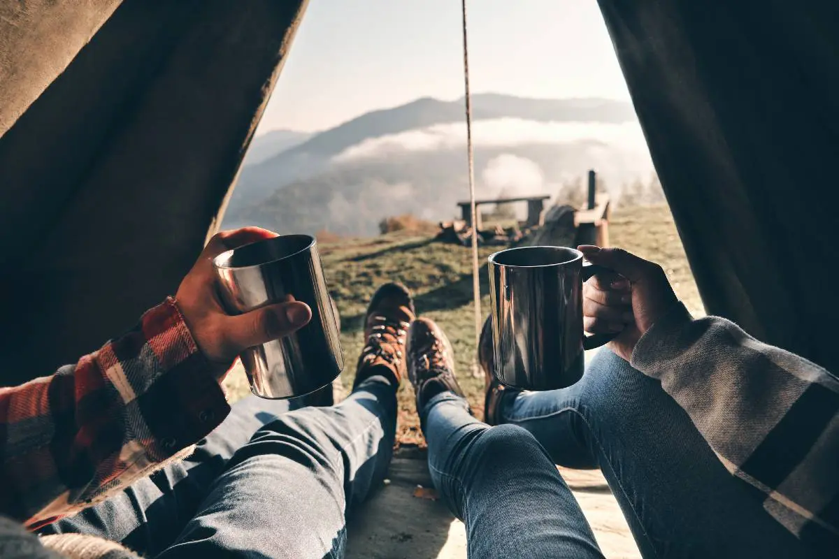 Two people lying inside a tent and holding up coffee mugs with a view of outside through their open tent, which is the most important item on a list of camping must haves
