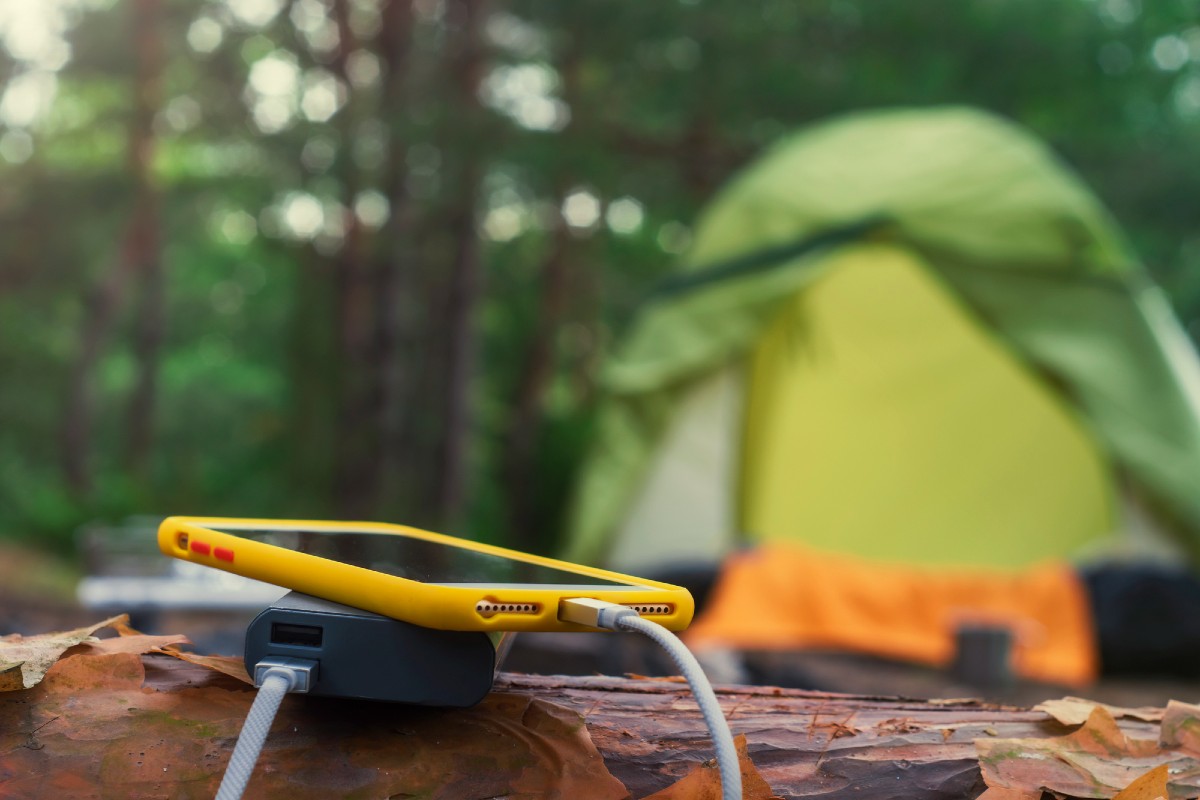 A tent in the background and a cell phone charging on a solar power bank, one of the most useful camping must haves