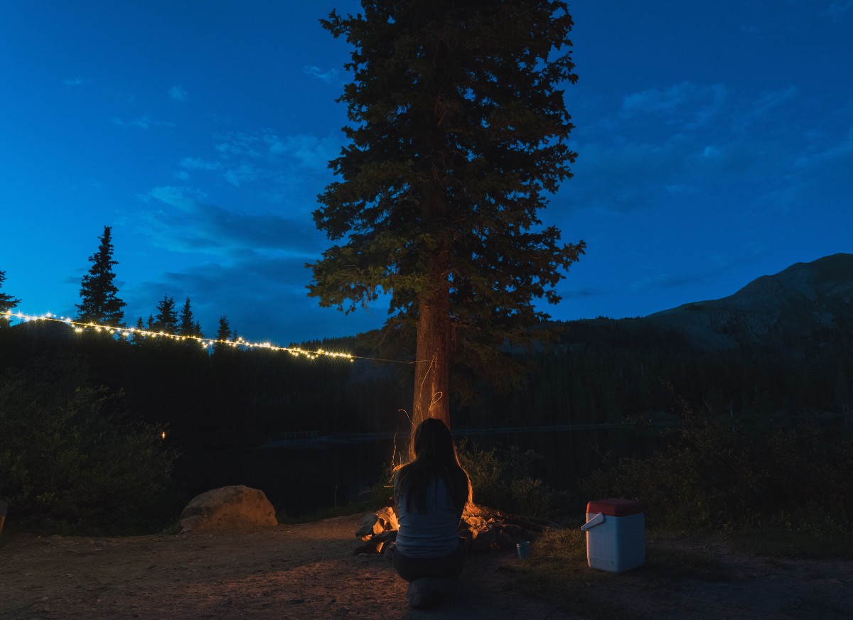 A woman sitting by a campfire near a tree with some top camping must haves, string lights, in the background