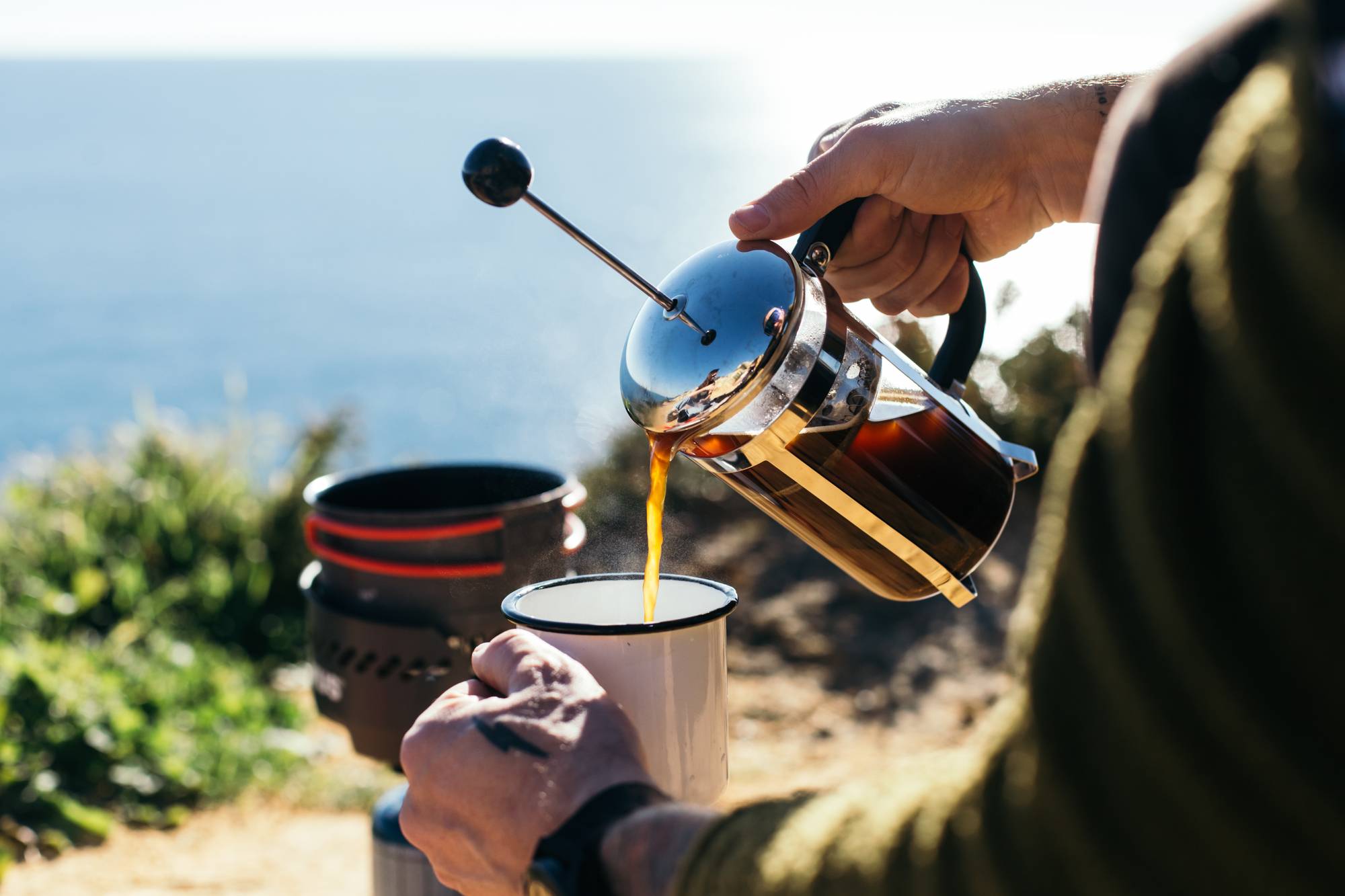 A man pouring cofee out of a camp French press into a mug near a body of water