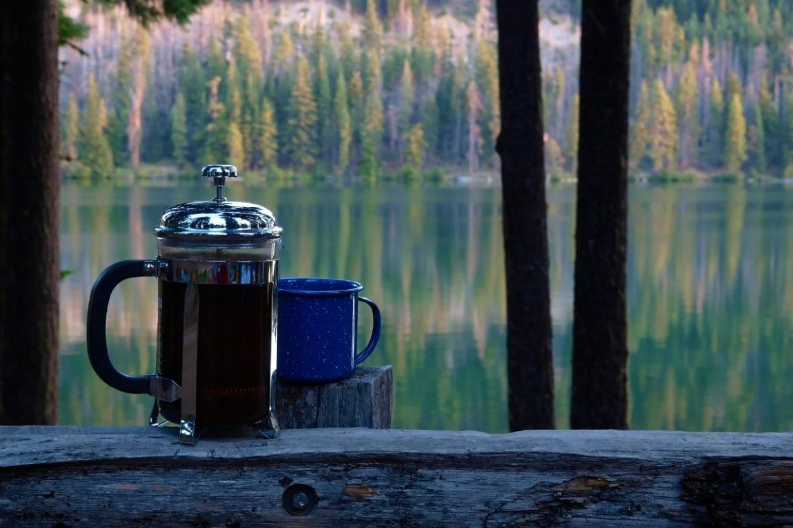 A camping French press next to a blue mug sitting on a ledge overlooking a mountain lake
