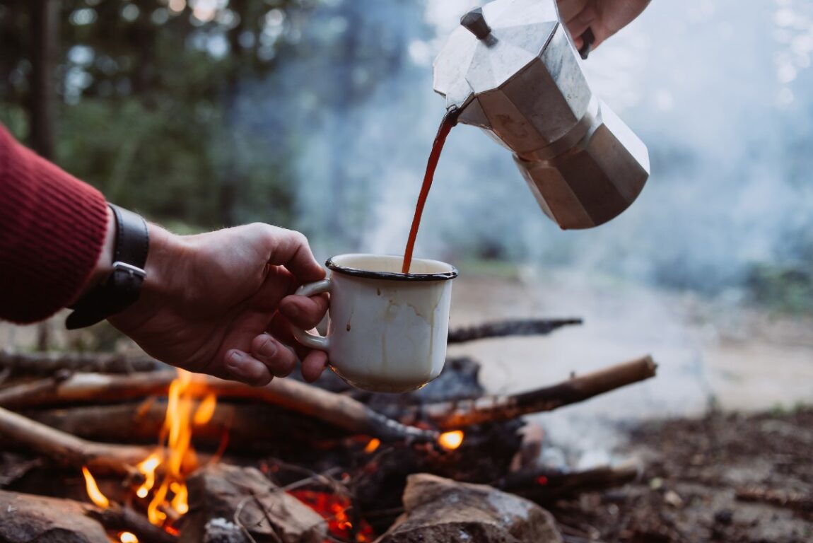 Someone pouring from a Bialetti camp percolator into a cup next to a campfire