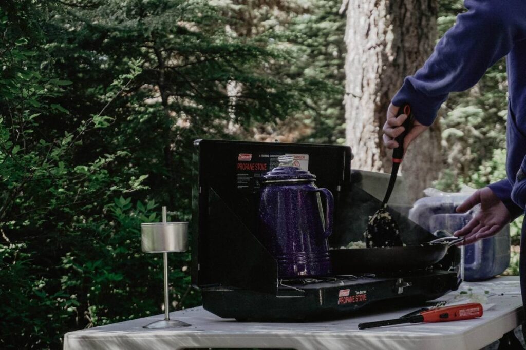 Someone cooking on a camp stove while a blue camping percolator makes coffee