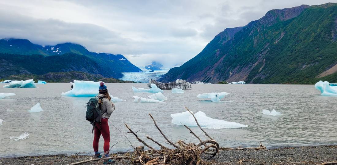 A woman wearing a hiking backpack standing at the shores of Grewingk Glacier Lake
