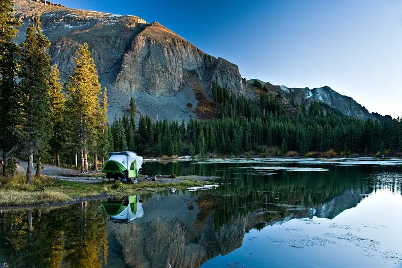 A green and white popup camper next to a lake in front of the mountains at one of the nicest campsites near Telluride