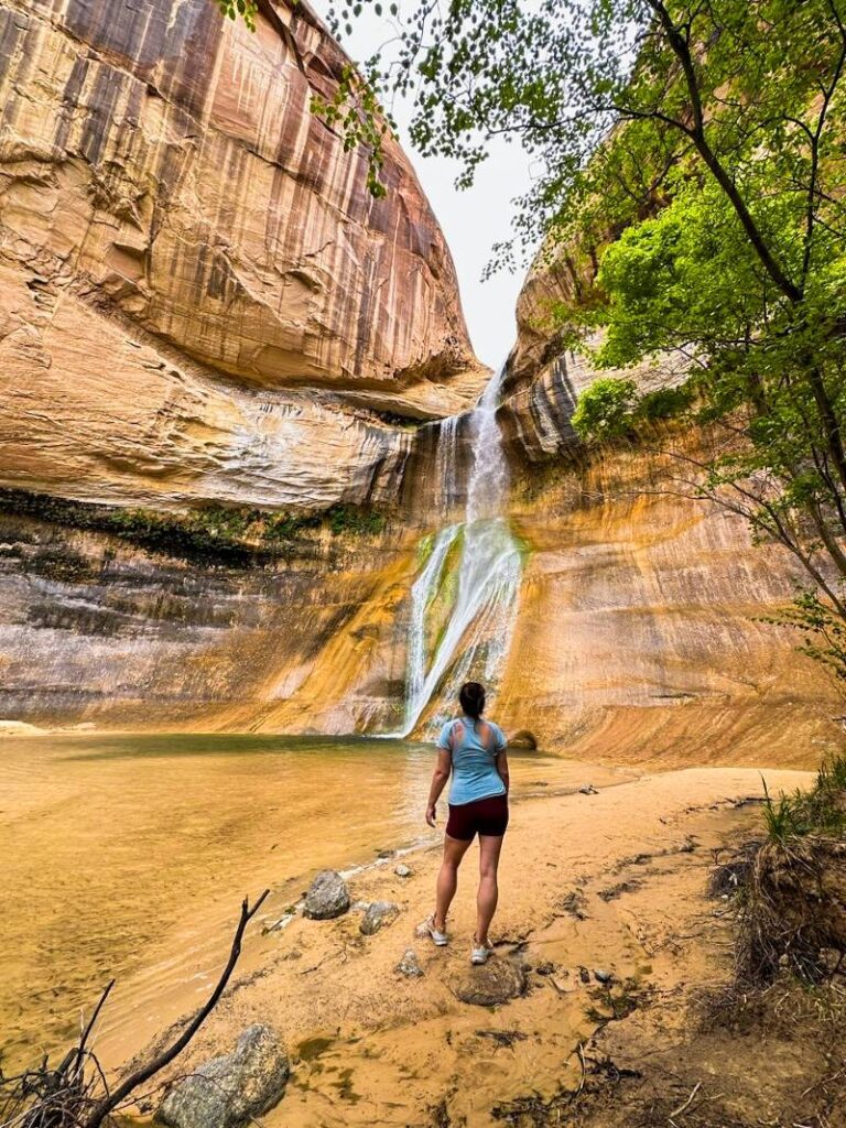 A woman wearing hiking gear and standing on the beach area in front of Lower Calf Creek Falls