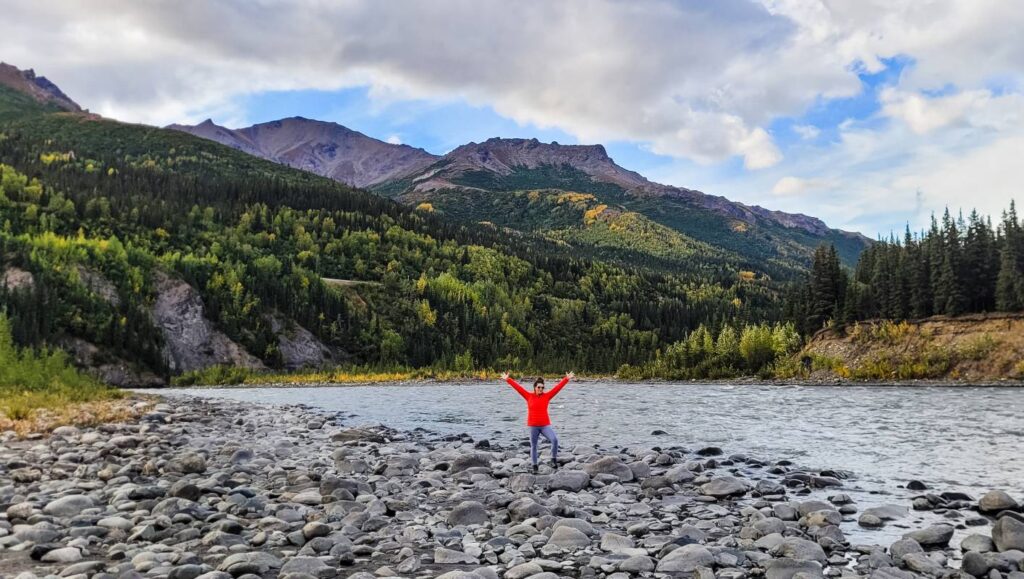 A woman throwing her hands out and wearing a bright red jacket and gray hiking leggings for women next to a river in Denali National Park